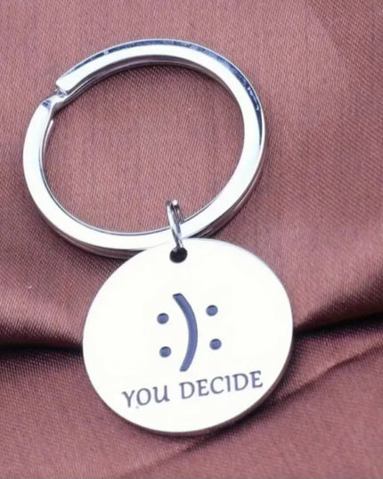 Silver keychain with a circular tag featuring a smiley face and 'YOU DECIDE' text on a brown fabric background.