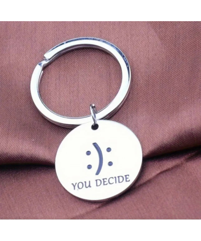 Silver keychain with a circular tag featuring a smiley face and 'YOU DECIDE' text on a brown fabric background.